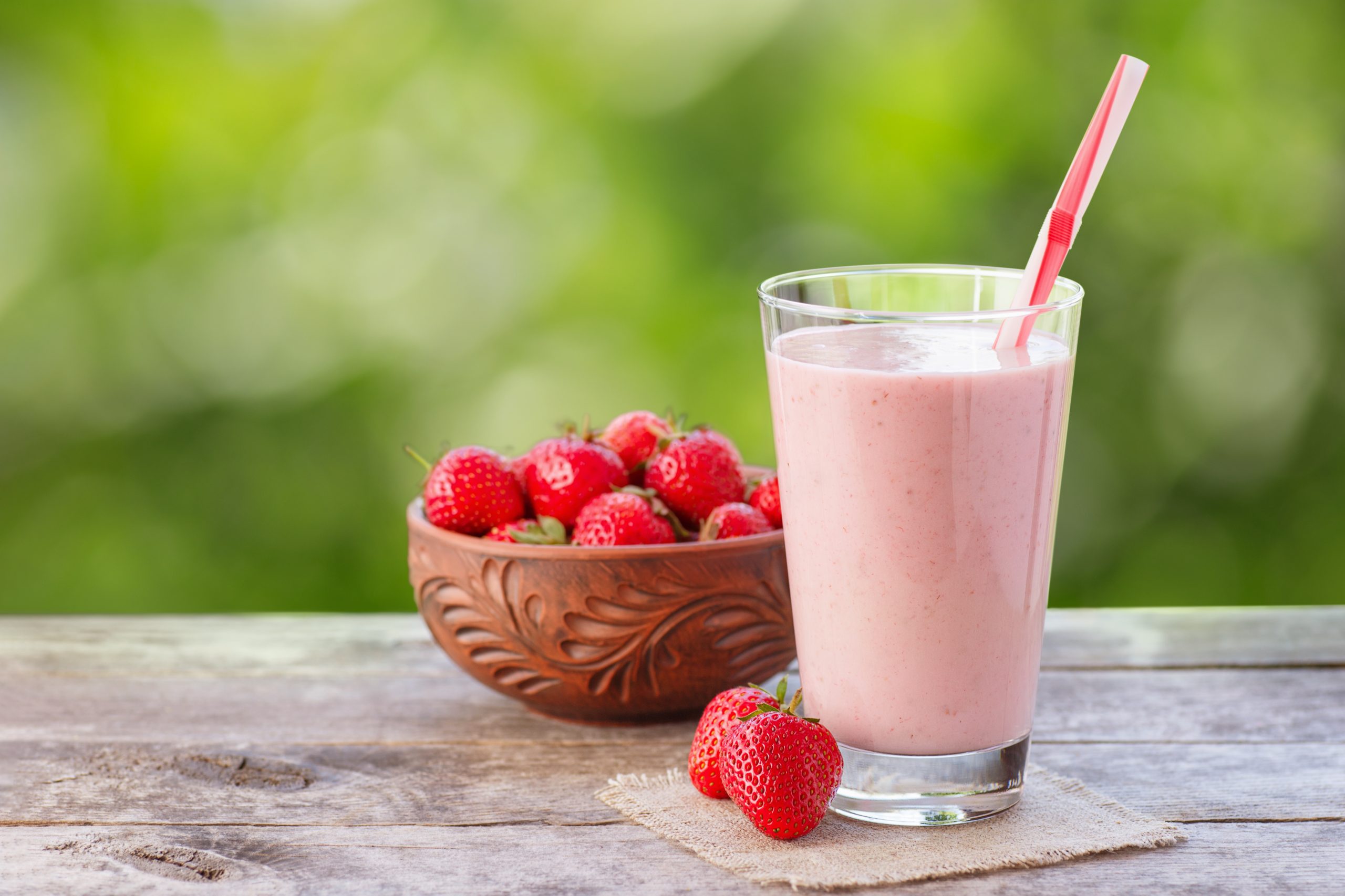 glass of strawberry milkshake and fresh ripe berries in bowl on wooden table with green blurred natural background